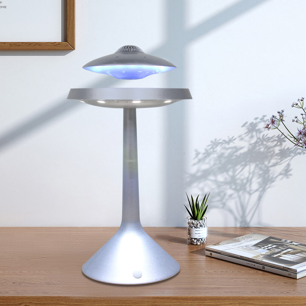 A Floating UFO Speaker with a modern design, emitting blue light and resembling a flying saucer, stands on a wooden desk next to a small potted plant and a book.