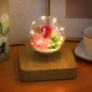A levitating glass orb with flowers and lights, set on a wooden base, next to a teacup on a table.